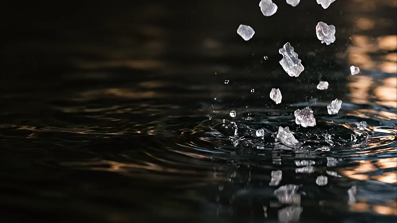Small mineral flakes falling into dark water, with fine ripples and silver reflections.