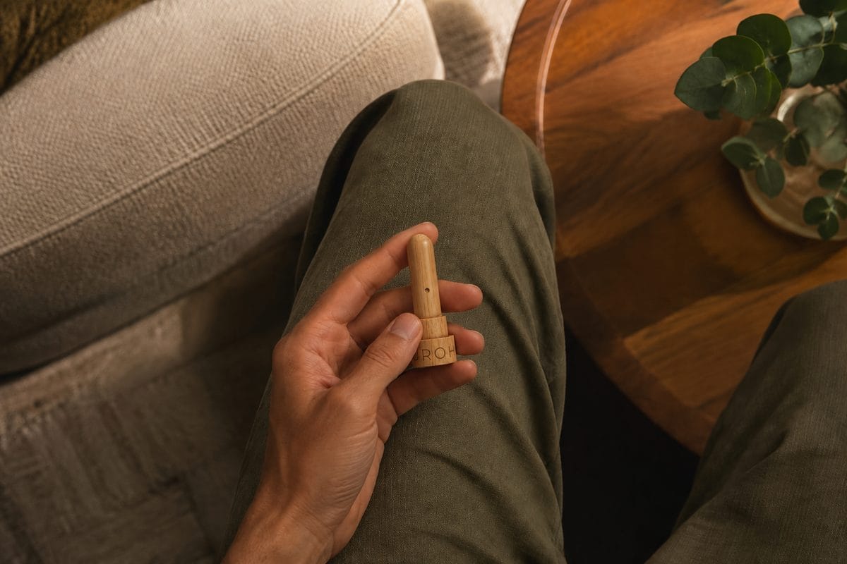 A hand holds an open bamboo Neurohaler with its rounded insert and small side opening above olive trousers.
